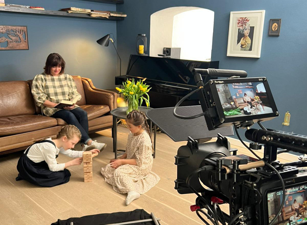 Im Titelbild unseres Imagefilms sieht man eine Frau und zwei Kinder, welche in einem Wohnzimmer sitzen. Die Frau liest ein Buch, die Kinder spielen Jenga. Im rechten Teil des Bildes sieht man eine Kamera, welche die Situation filmt.