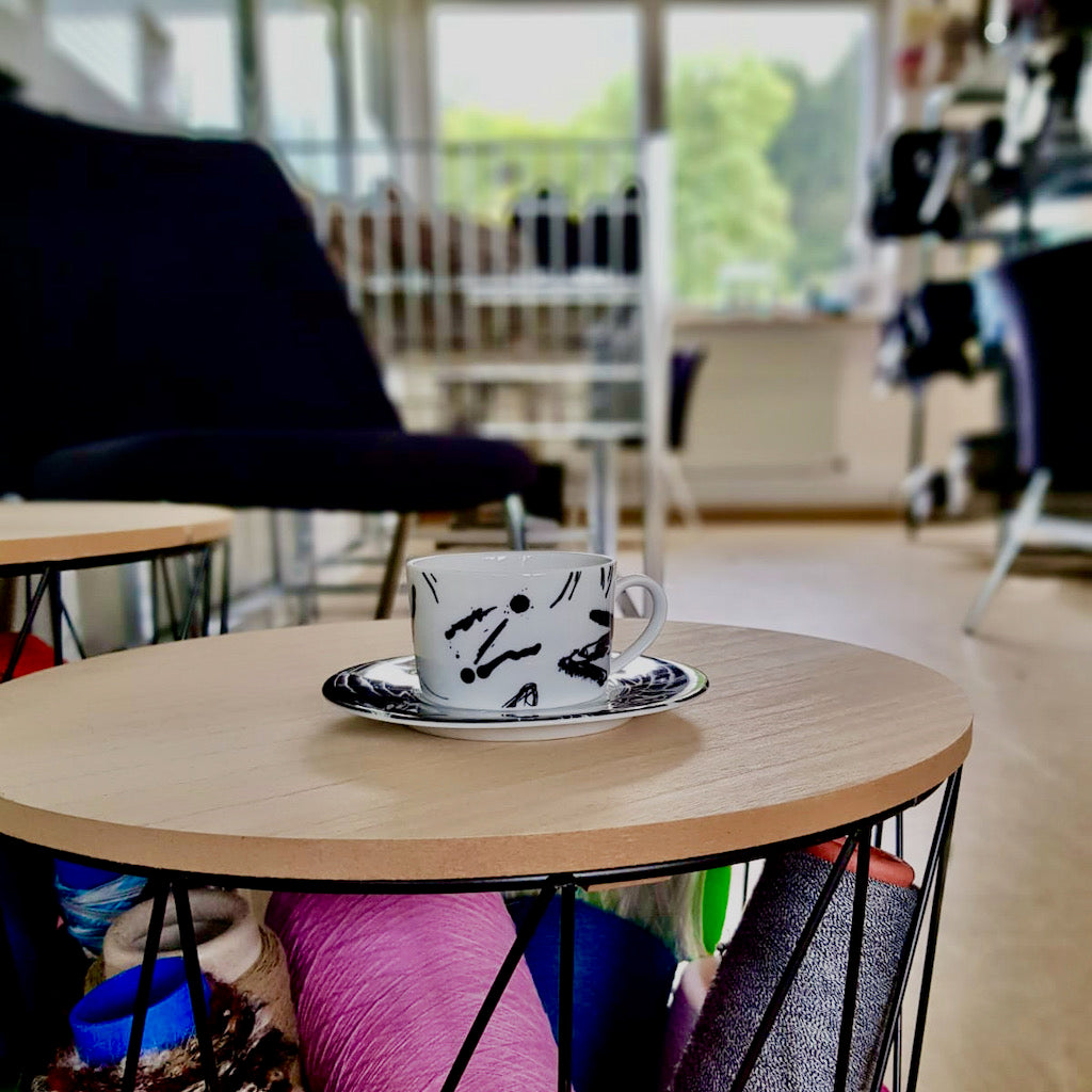 Kaffeetasse auf einem mit Garnspulen gefülltem Beistelltisch. Im Hintergrund ist verschwommen unser Werksverkauf erkennbar.